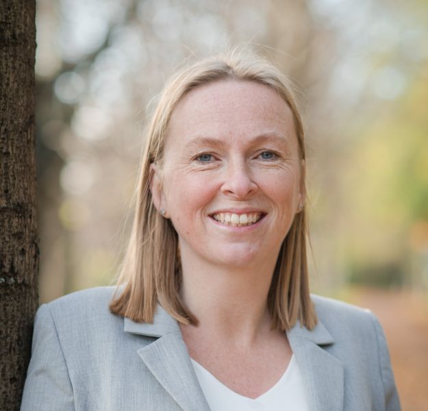 Portrait of a smiling woman with shoulder-length blonde hair, wearing a light grey blazer and white top, standing beside a tree outdoors with softly blurred trees in the background.