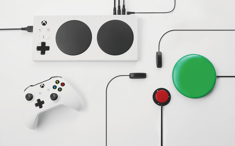 A picture with a standard xbox controller sitting just below the Xbox adaptive controller. The adaptive controller has 4 switches attached to it of various colours. 